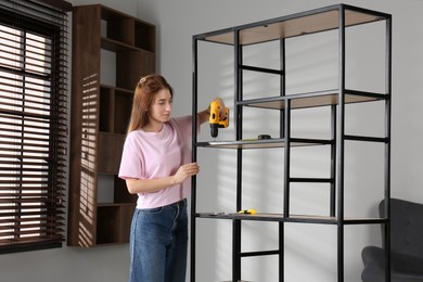 Photo of Woman with electric screwdriver assembling shelving unit in room