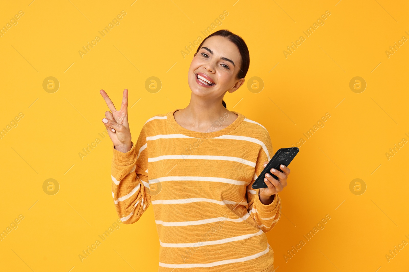 Photo of Happy woman with smartphone showing peace sign on yellow background