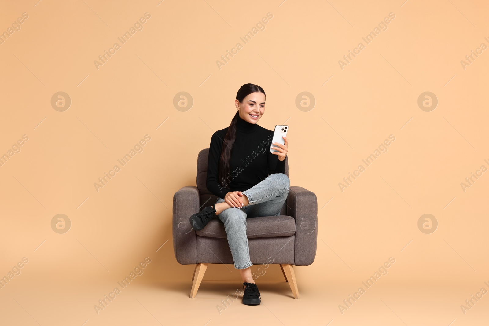 Photo of Smiling woman with smartphone sitting on armchair against beige background