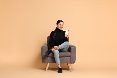 Photo of Smiling woman with smartphone sitting on armchair against beige background