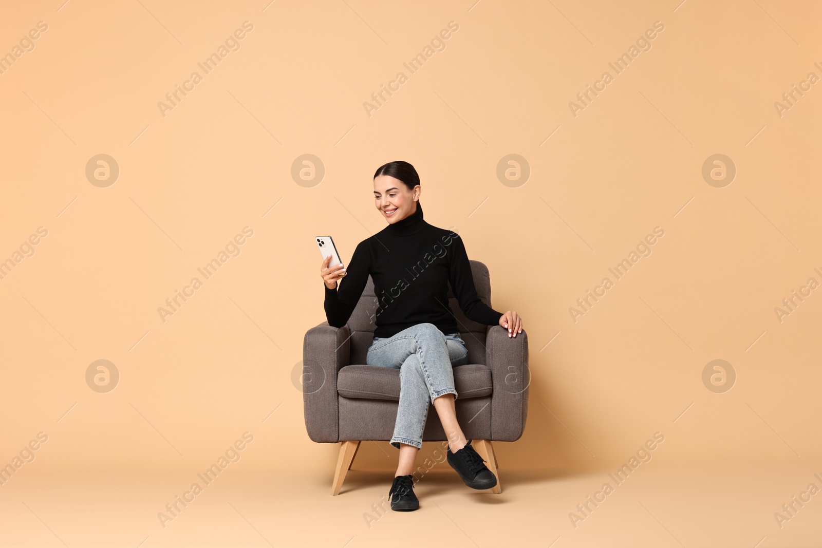 Photo of Smiling woman with smartphone sitting on armchair against beige background