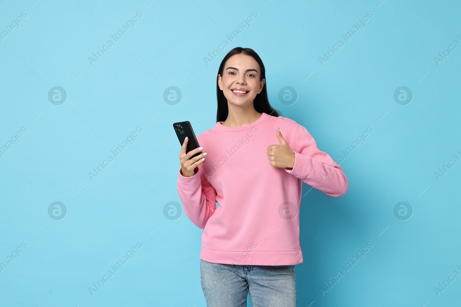 Photo of Portrait of smiling woman with smartphone showing thumbs up on light blue background