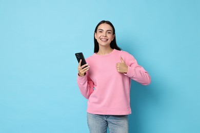 Portrait of smiling woman with smartphone showing thumbs up on light blue background