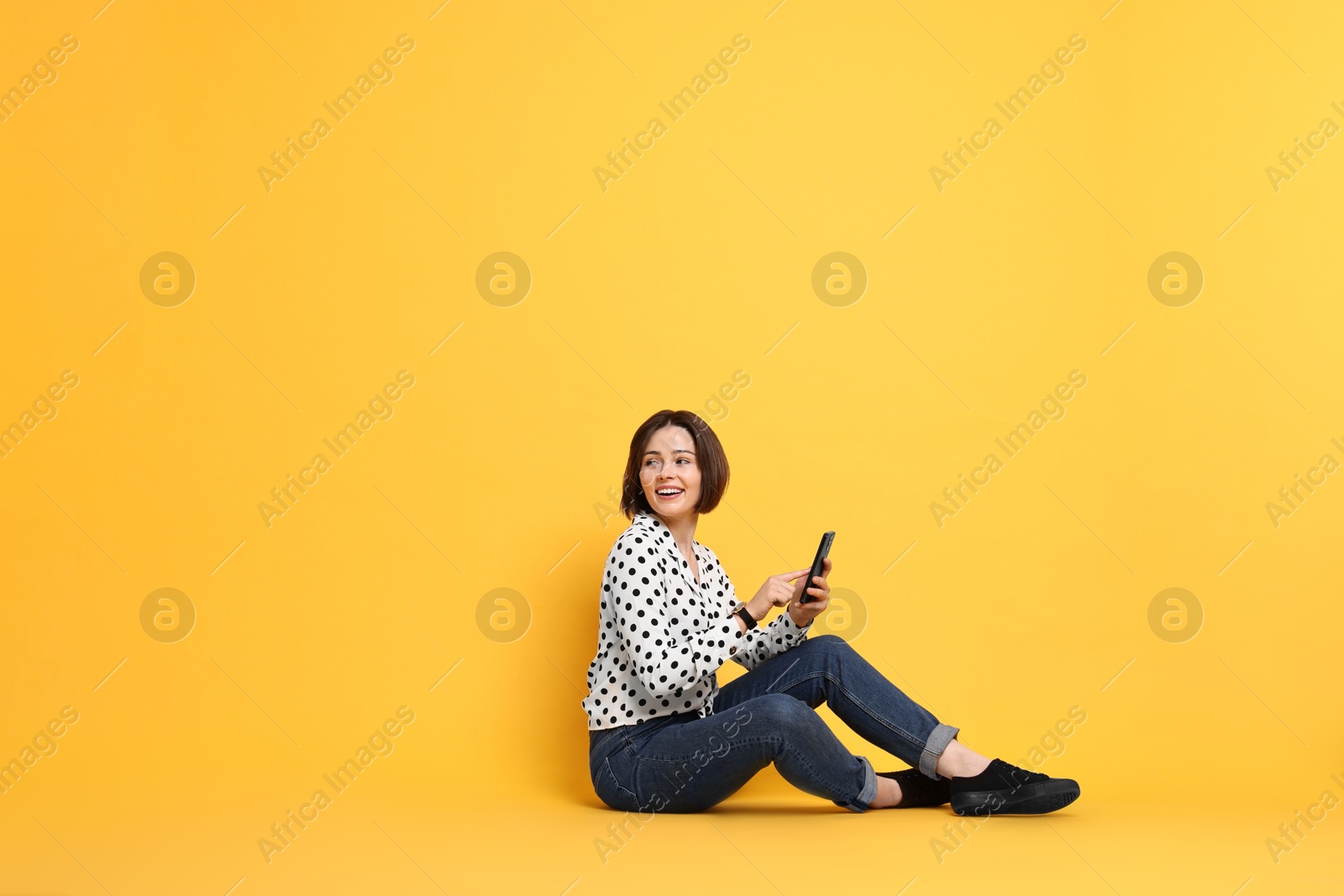 Photo of Smiling woman with smartphone sitting on yellow background. Space for text