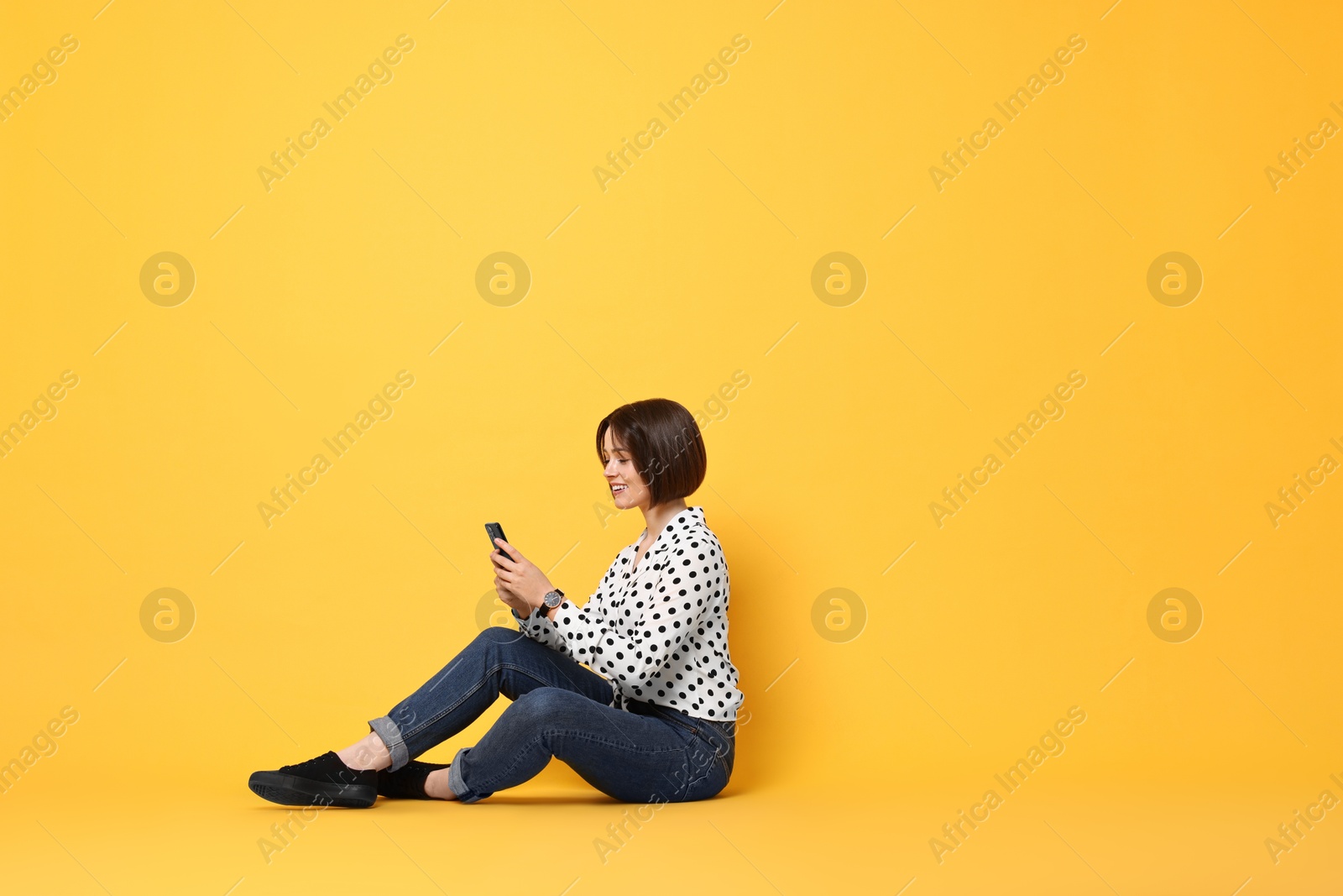 Photo of Smiling woman with smartphone sitting on yellow background. Space for text