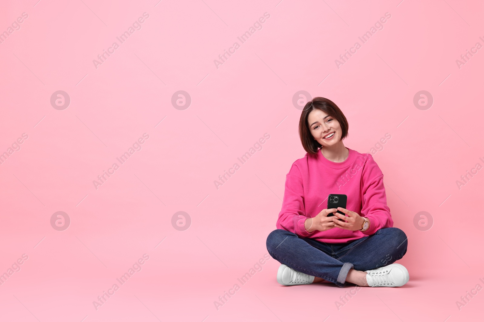 Photo of Smiling woman with smartphone sitting on pink background. Space for text