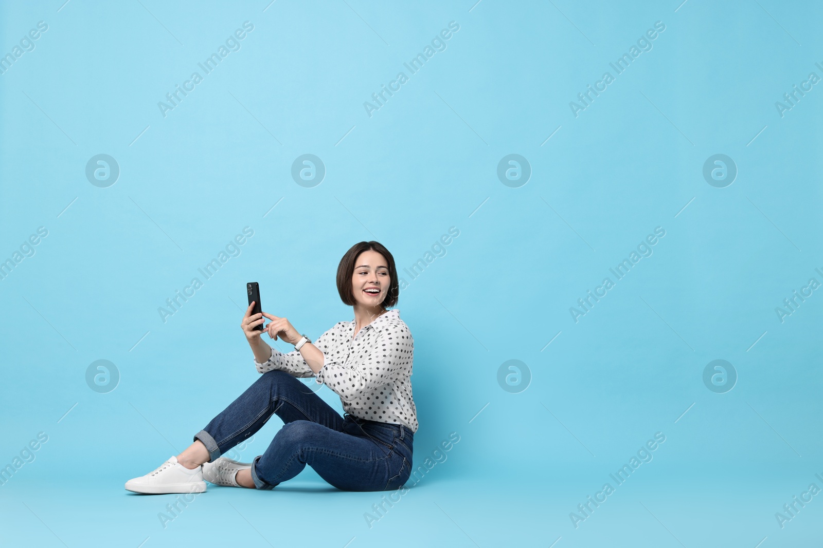 Photo of Smiling woman with smartphone sitting on light blue background. Space for text