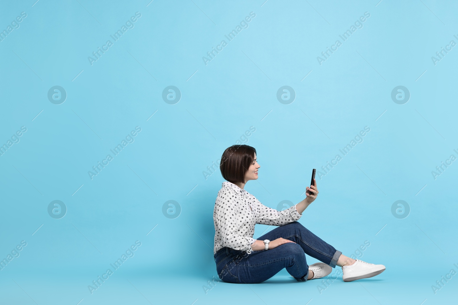 Photo of Smiling woman with smartphone sitting on light blue background. Space for text