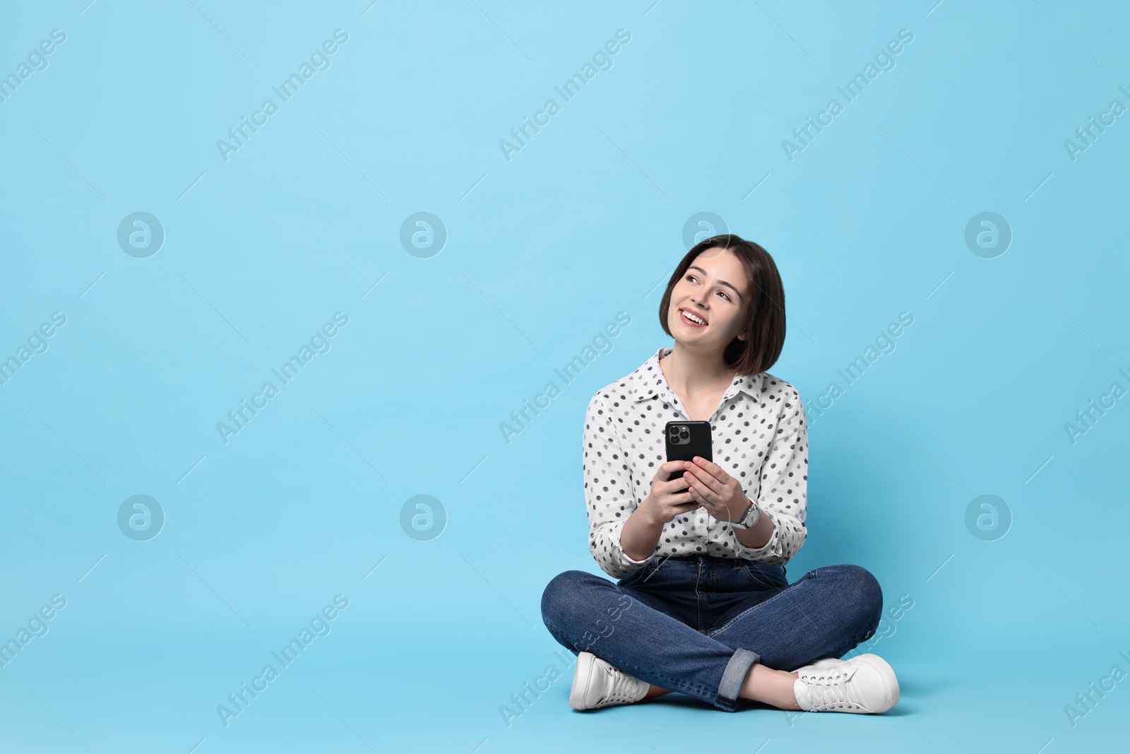 Photo of Smiling woman with smartphone sitting on light blue background. Space for text