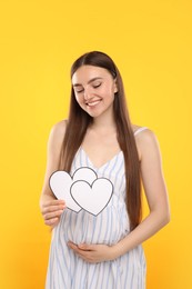 Photo of Expecting twins. Pregnant woman holding two paper cutouts of hearts on yellow background