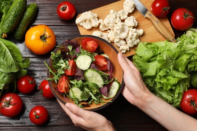 Photo of Healthy vegetarian food. Woman holding bowl of salad at wooden table with vegetables, top view