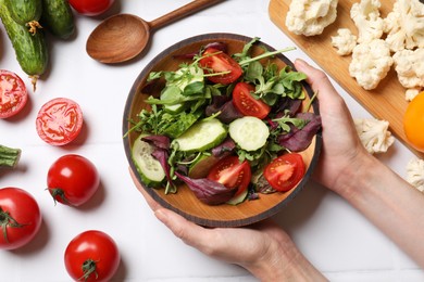 Photo of Healthy vegetarian food. Woman holding bowl of salad at white tiled table with vegetables, top view