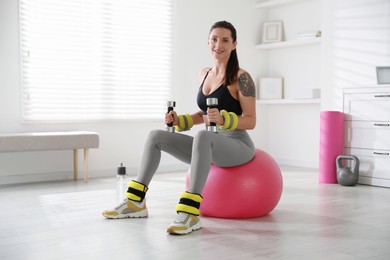 Beautiful woman with ankle weights and dumbbells training on fitness ball indoors