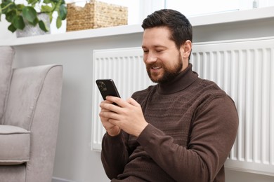 Photo of Happy man using smartphone near heating radiator at home