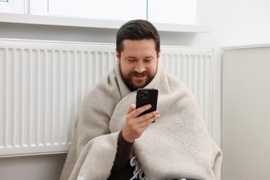 Photo of Happy man using smartphone near heating radiator at home