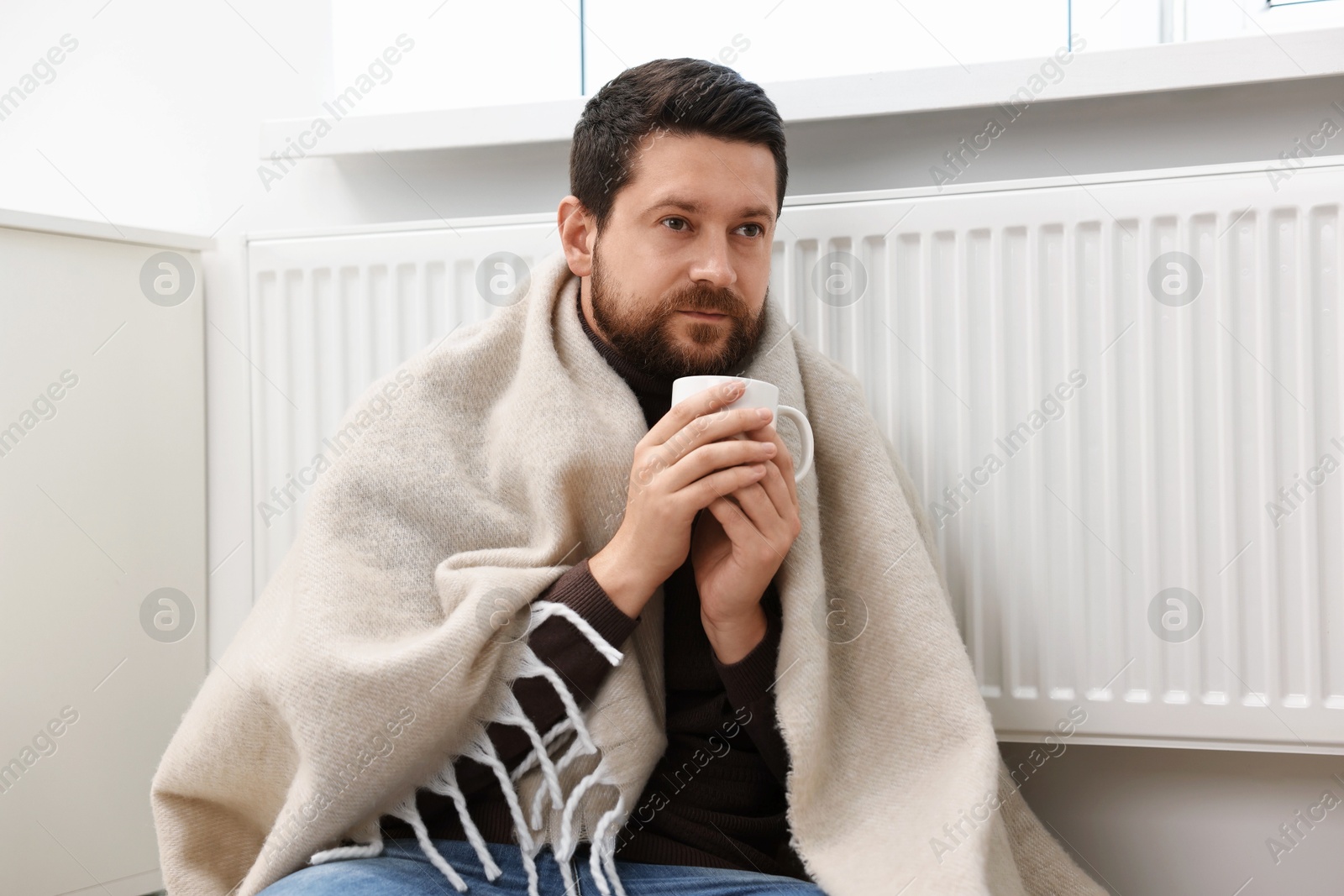 Photo of Man with cup of hot drink near heating radiator at home