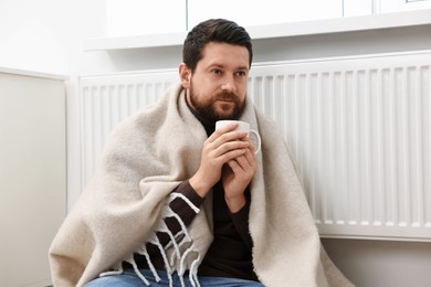 Man with cup of hot drink near heating radiator at home