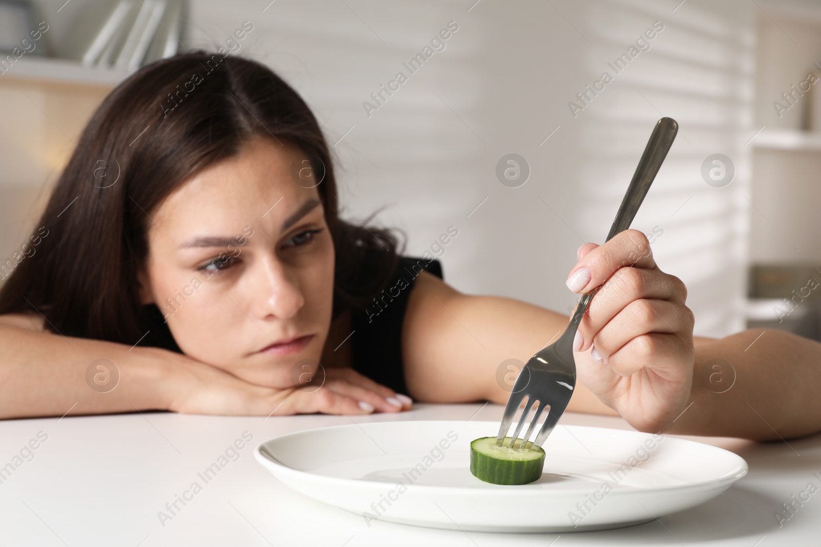 Photo of Eating disorder. Sad woman holding fork with cucumber at table indoors