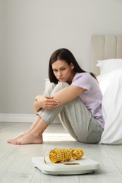 Eating disorder. Sad woman sitting near scale and measuring tape on floor indoors