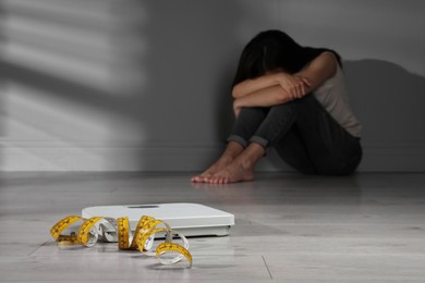 Eating disorder. Depressed woman sitting near wall indoors, focus on scale and measuring tape