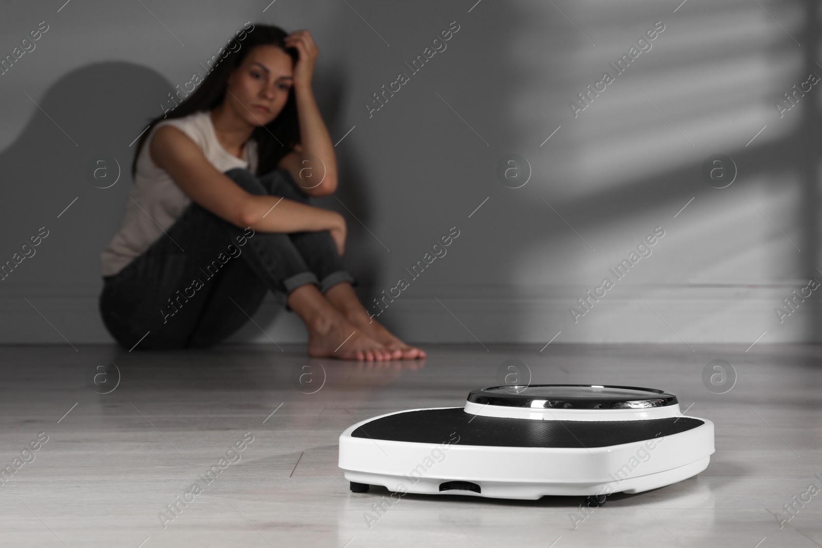 Photo of Eating disorder. Sad woman sitting near wall indoors, focus on scale