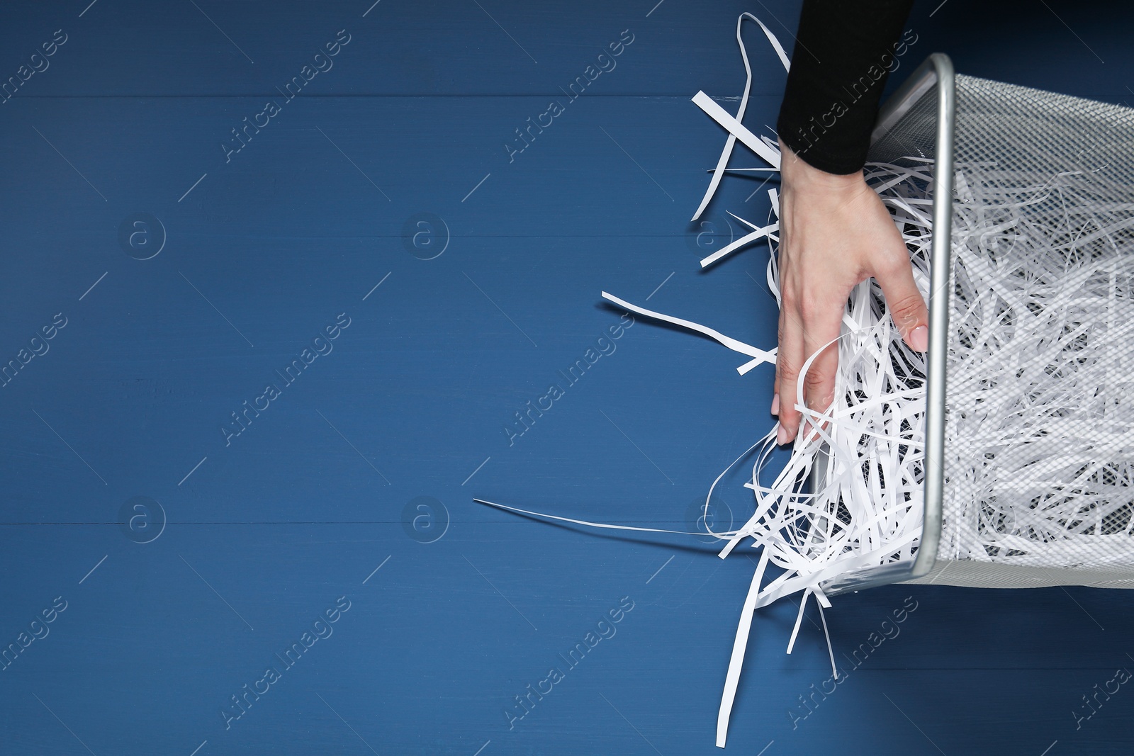 Photo of Woman putting shredded paper strips into trash bin at blue wooden table, top view. Space for text