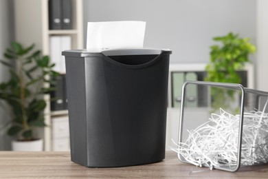 Photo of Destroying sheets with shredder and paper strips in metal bin on wooden table in office