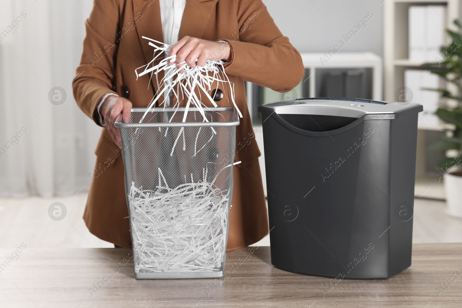 Photo of Woman putting shredded paper strips into trash bin at wooden table in office, closeup