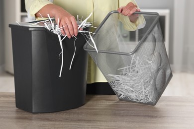 Photo of Woman putting shredded paper strips into trash bin at wooden table in office, closeup