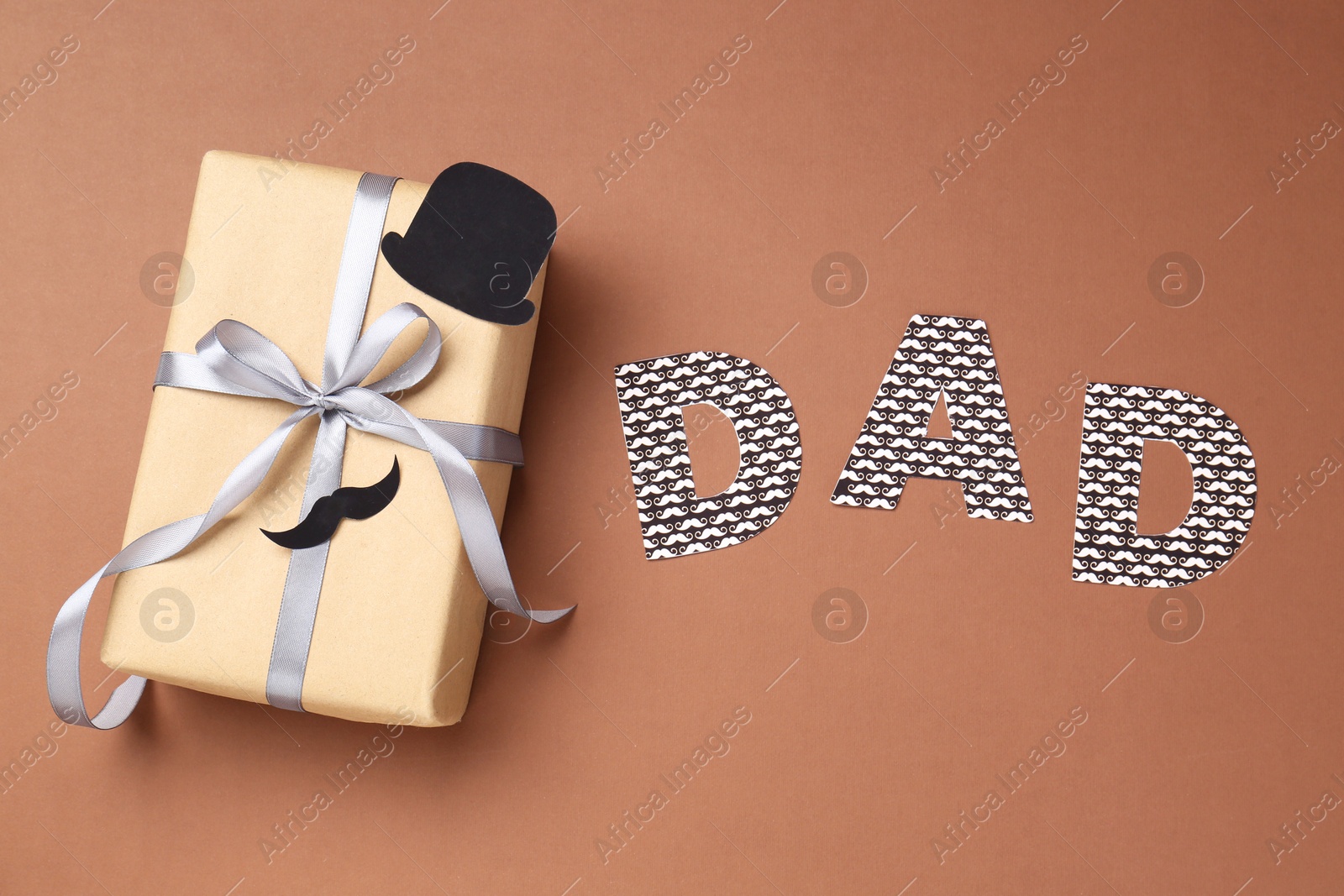 Photo of Happy Father's Day. Gift box with paper hat, moustache and word Dad on brown background, top view