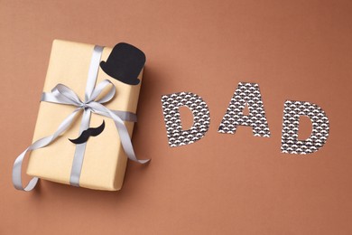 Photo of Happy Father's Day. Gift box with paper hat, moustache and word Dad on brown background, top view