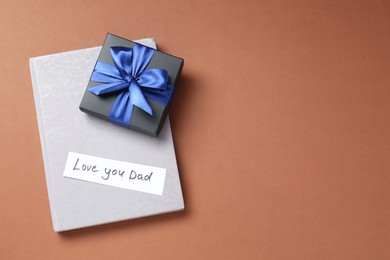 Happy Father's Day. Note with phrase Love You Dad, gift box and book on brown background, top view