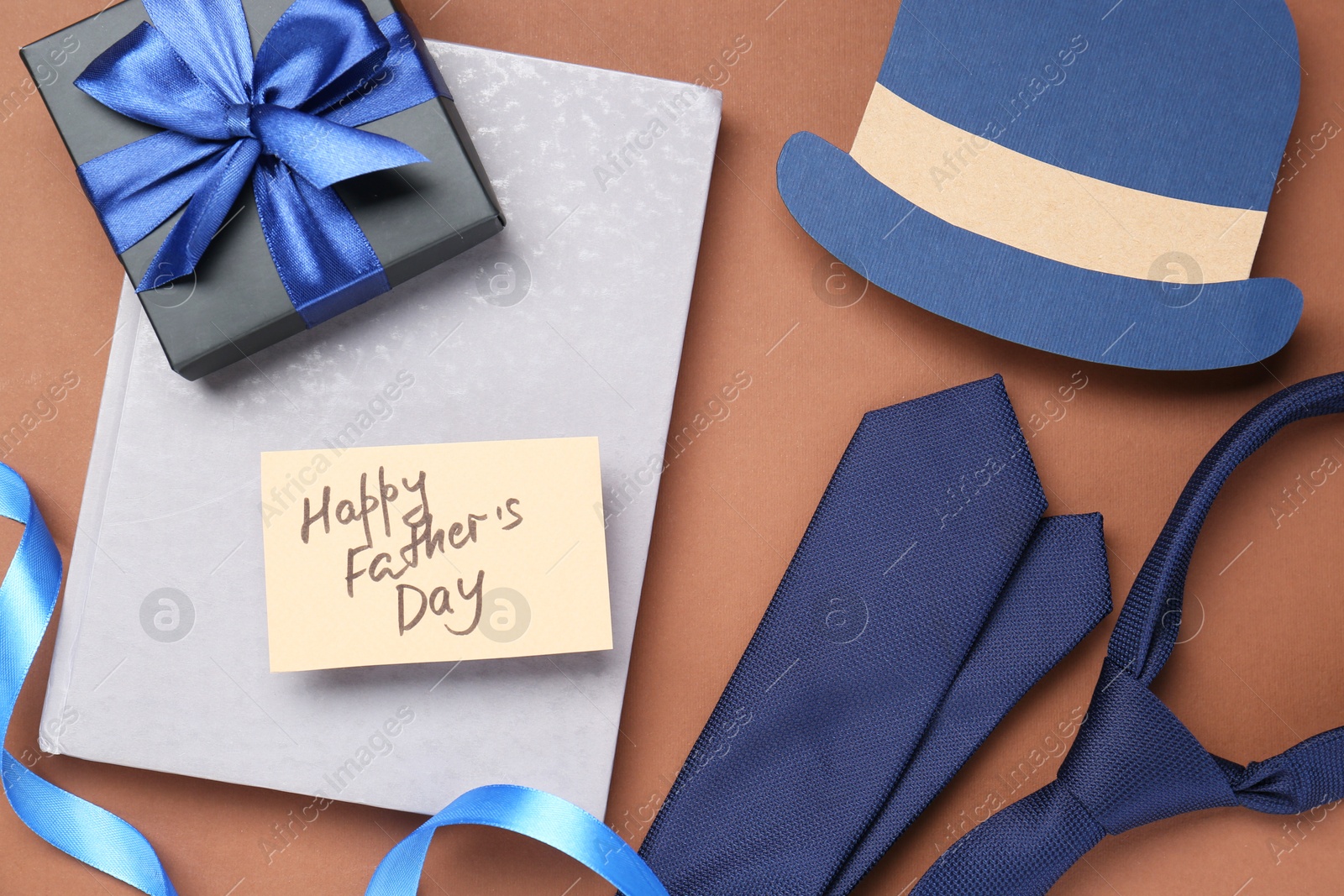 Photo of Card with phrase Happy Father's Day, gift, book, tie and paper hat on brown background, flat lay