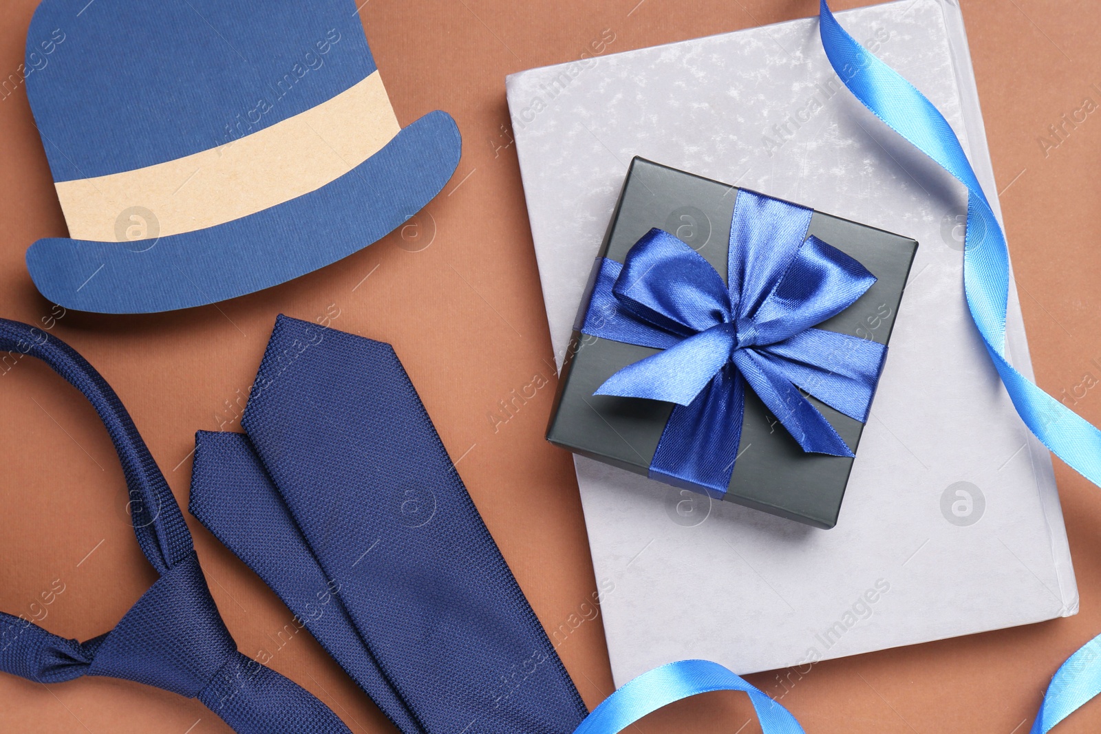 Photo of Happy Father's Day. Gift, book, tie and paper hat on brown background, flat lay