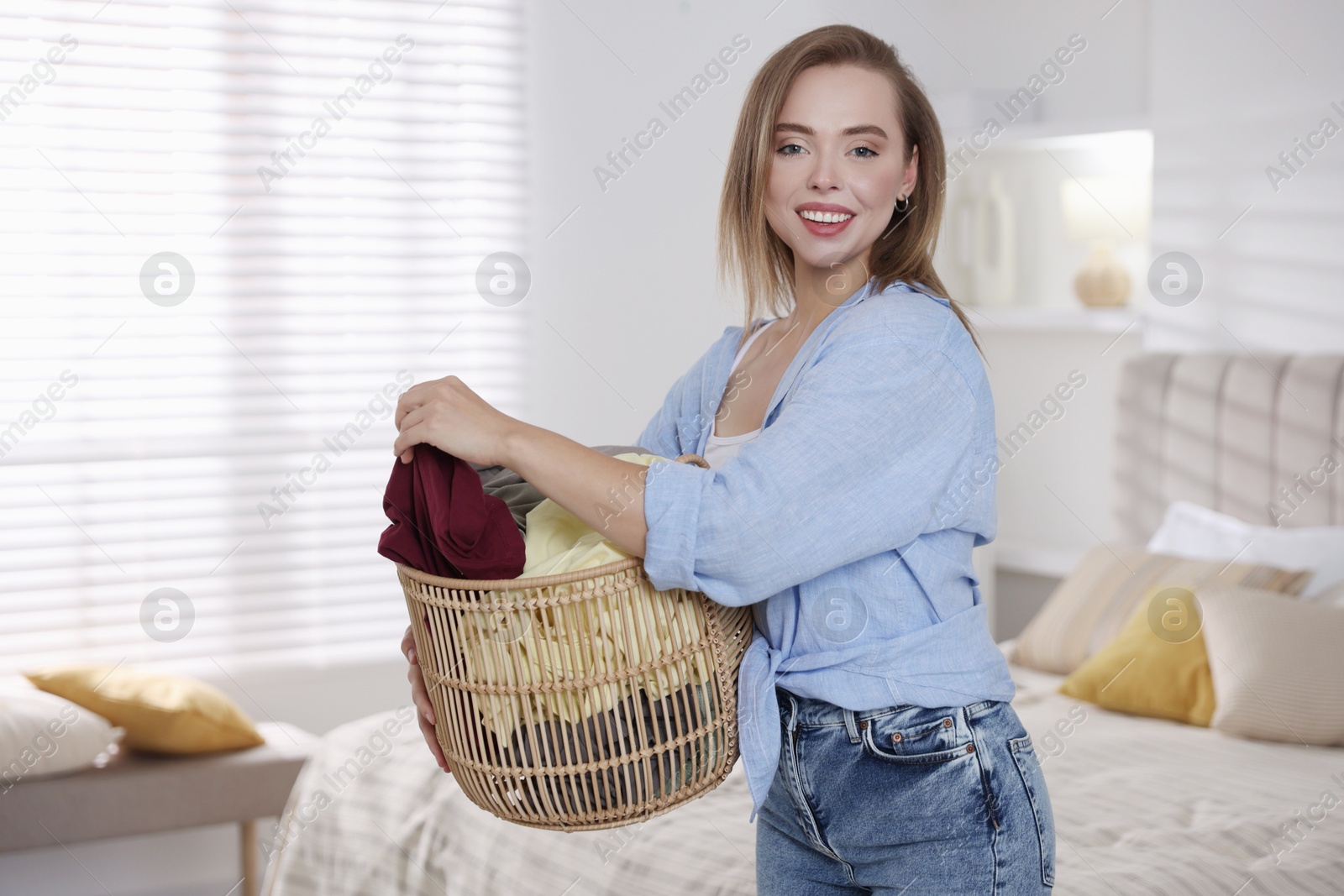 Photo of Happy young housewife with basket full of laundry at home