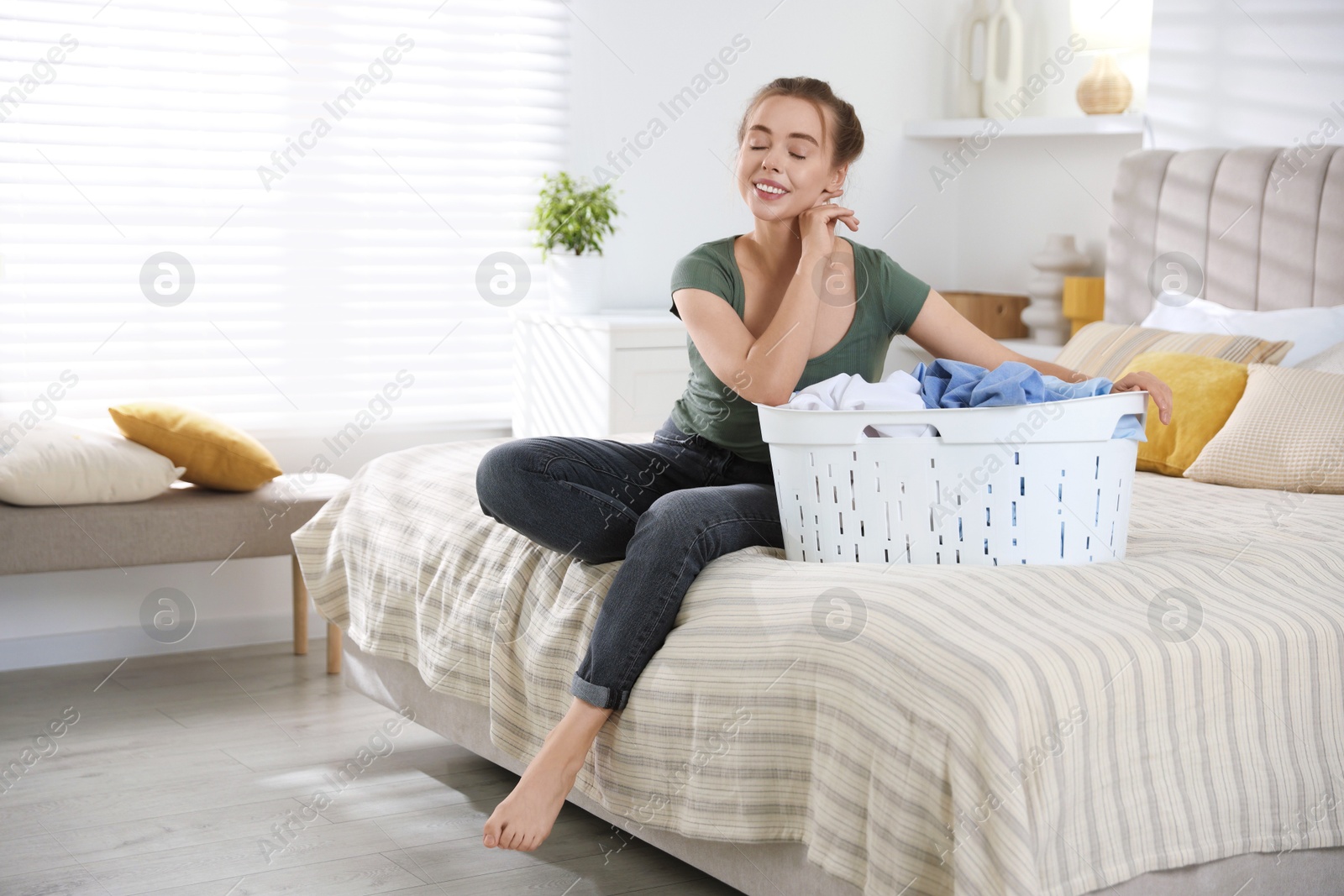 Photo of Happy young housewife with basket full of laundry on bed at home