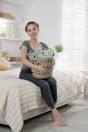 Happy young housewife with basket full of laundry on bed at home