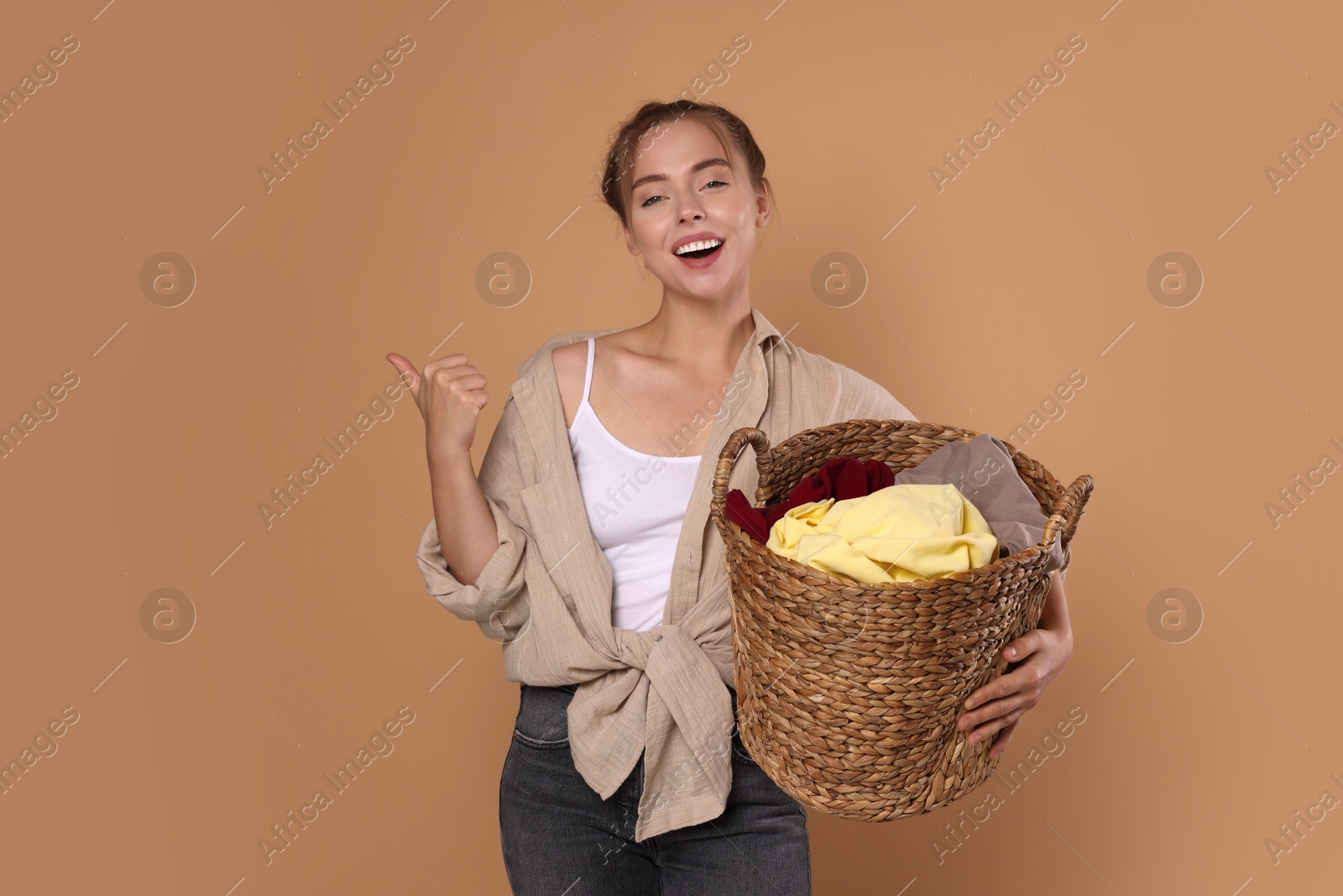 Photo of Happy young housewife with basket full of laundry showing thumbs up on pale orange background