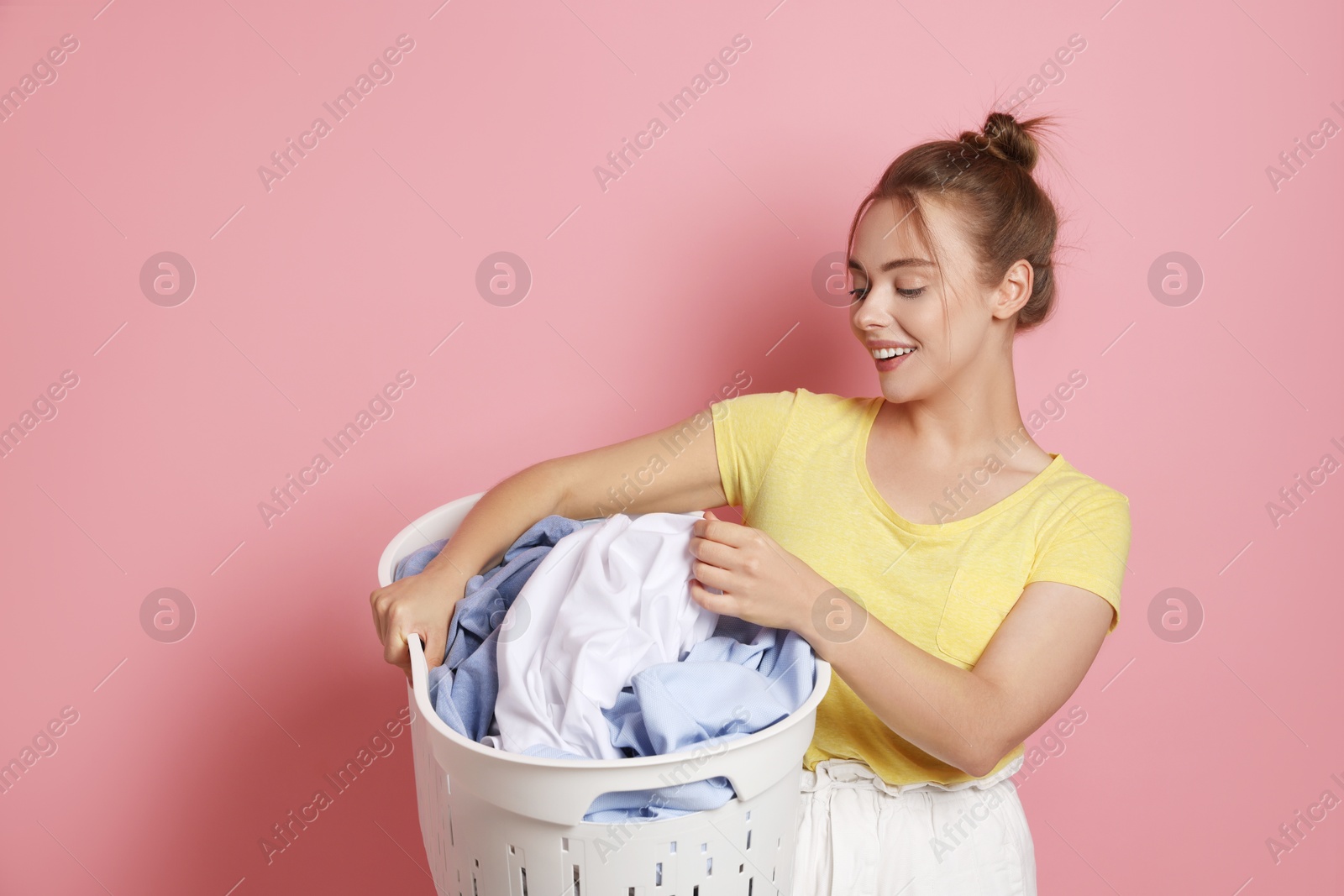 Photo of Happy young housewife with basket full of laundry on pale pink background. Space for text