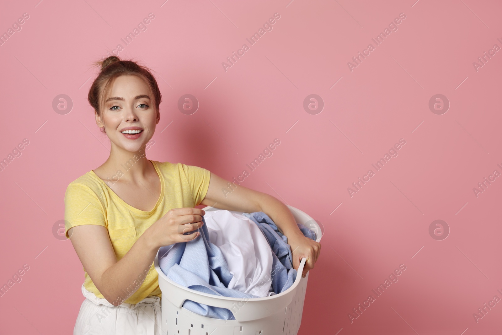 Photo of Happy young housewife with basket full of laundry on pale pink background. Space for text