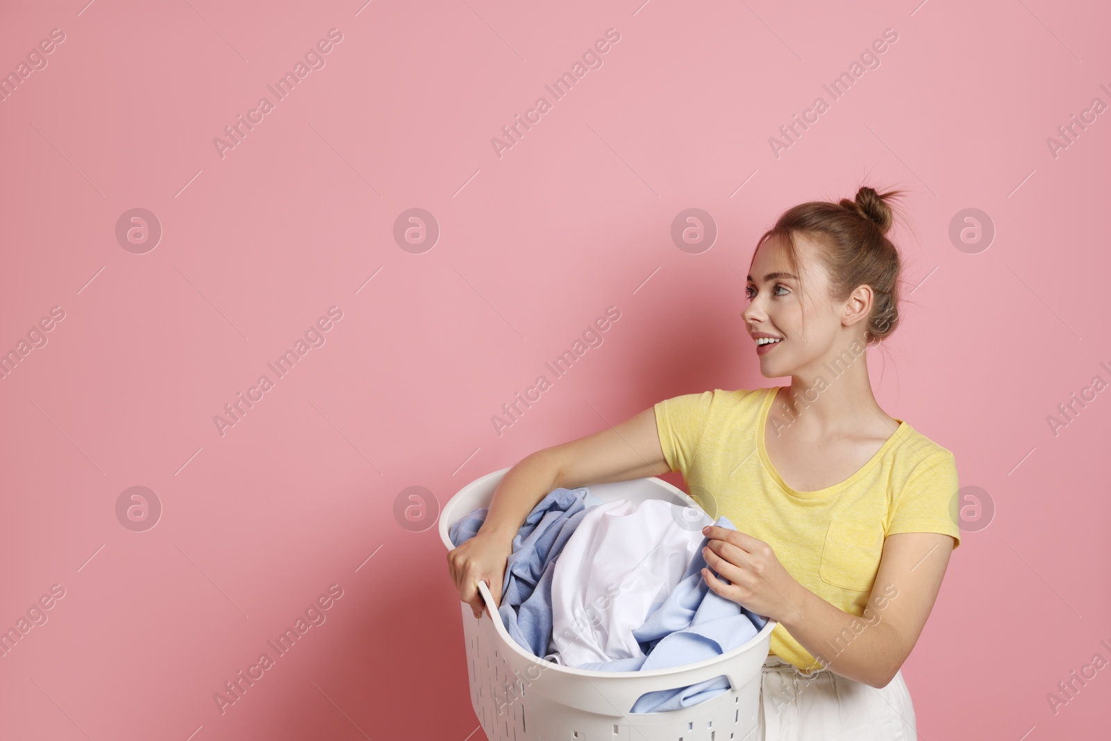 Photo of Happy young housewife with basket full of laundry on pale pink background. Space for text