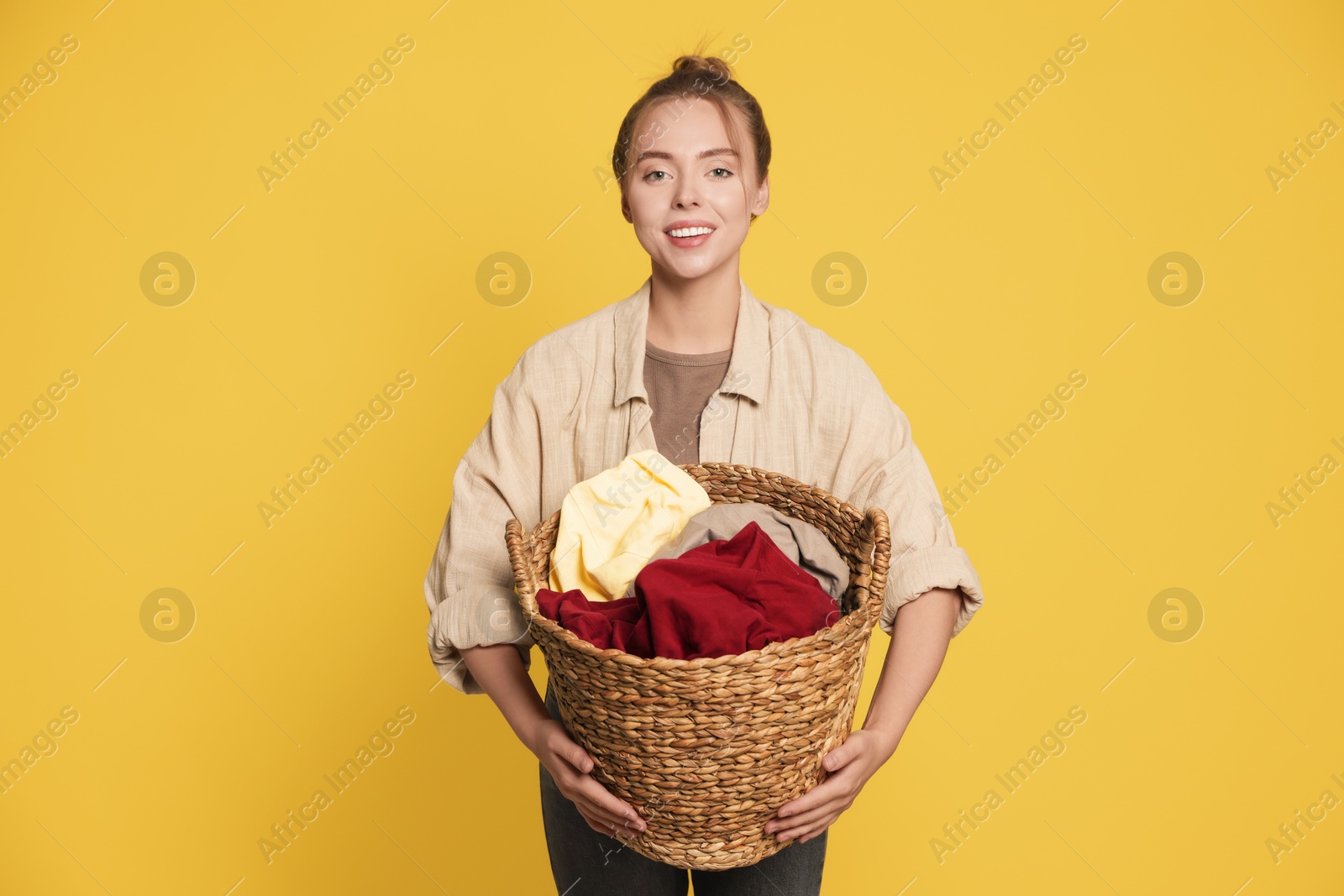 Photo of Happy young housewife with basket full of laundry on yellow background