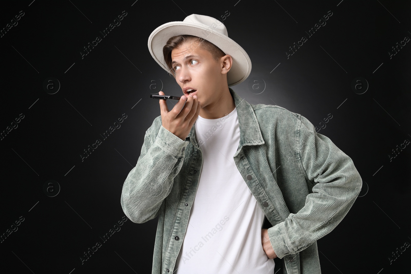 Photo of Young man recording voice message via smartphone on dark background