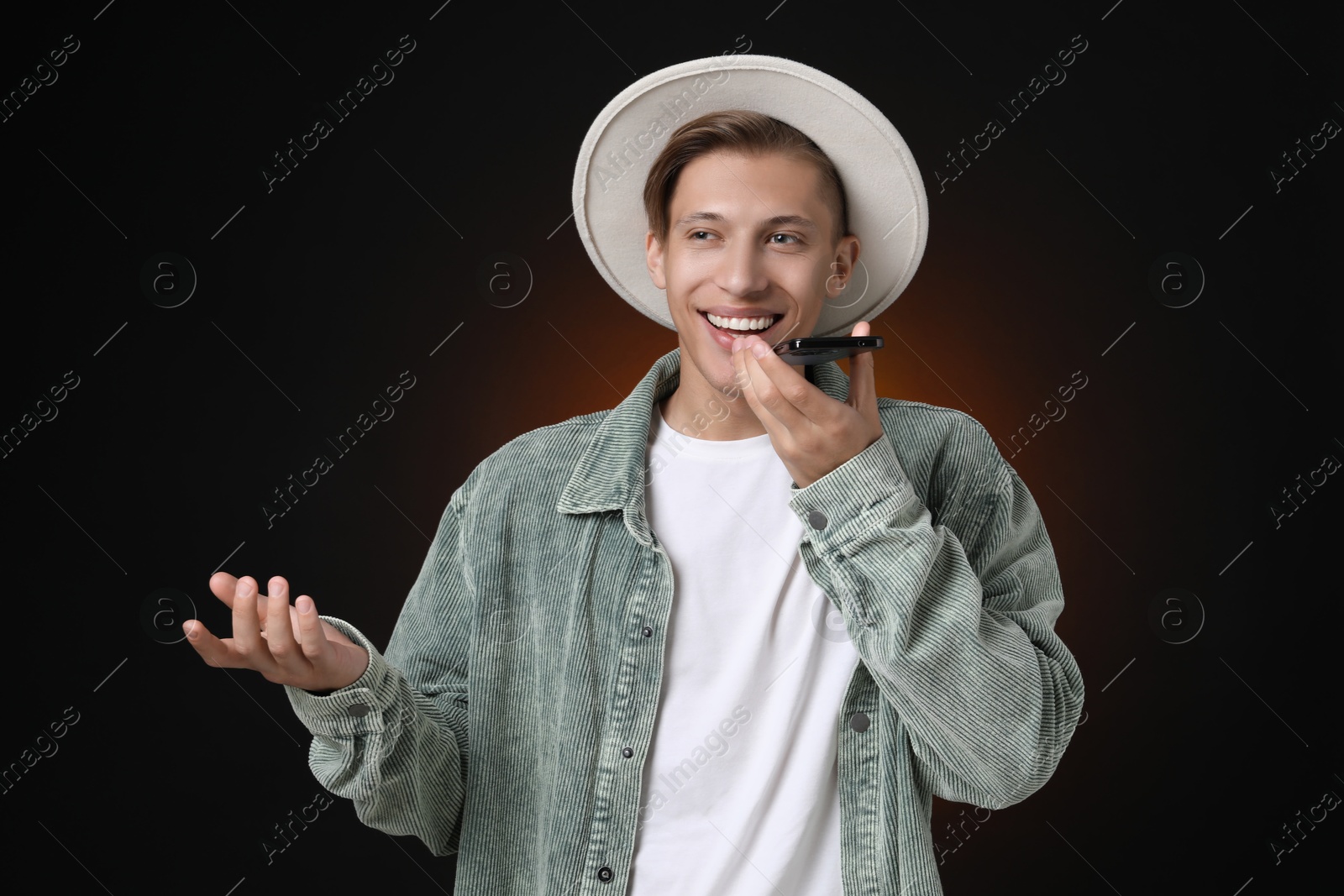 Photo of Young man recording voice message via smartphone on dark background