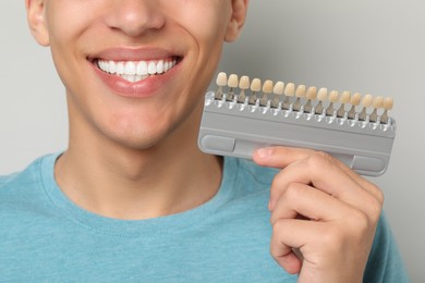 Happy young man with teeth color samples on gray background, closeup. Dental veneers