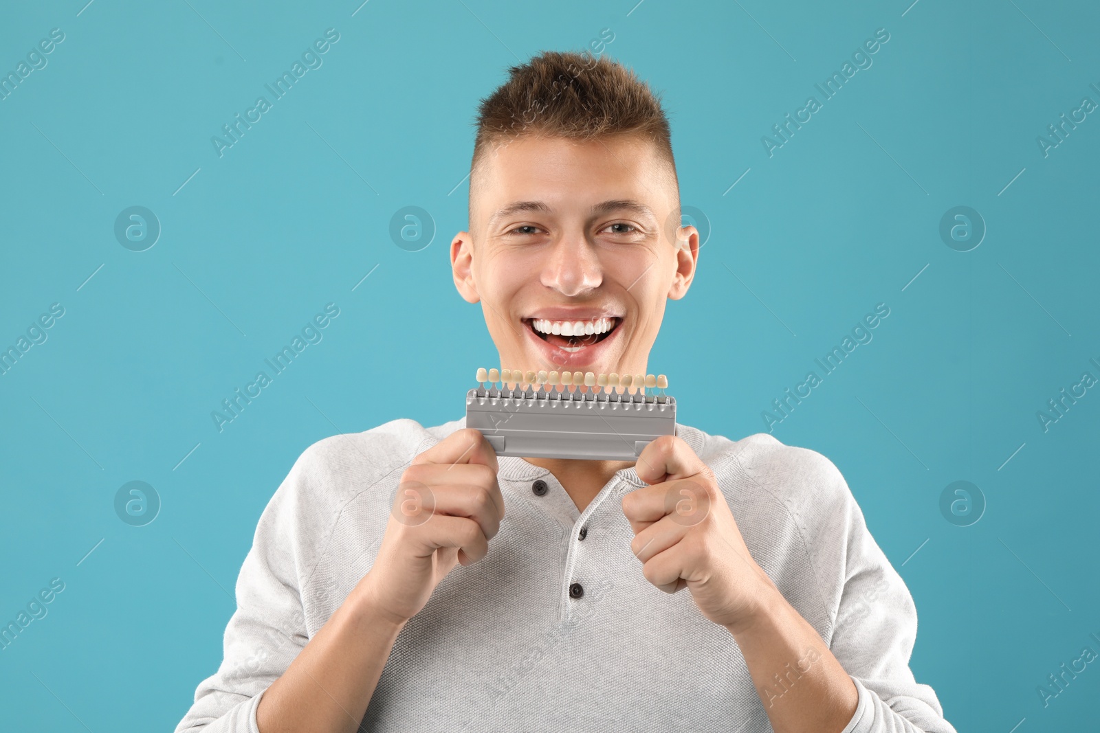 Photo of Happy young man with teeth color samples on light blue background. Dental veneers