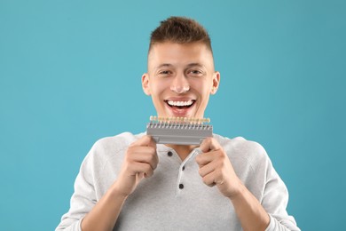 Photo of Happy young man with teeth color samples on light blue background. Dental veneers