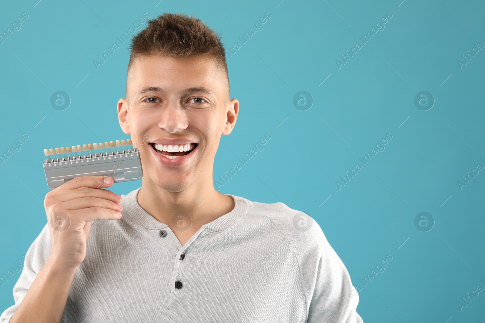 Photo of Happy young man with teeth color samples on light blue background, space for text. Dental veneers