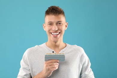 Photo of Happy young man with teeth color samples on light blue background. Dental veneers