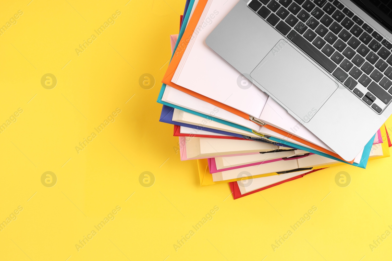 Photo of Stack of colorful books and laptop on yellow background, top view. Space for text